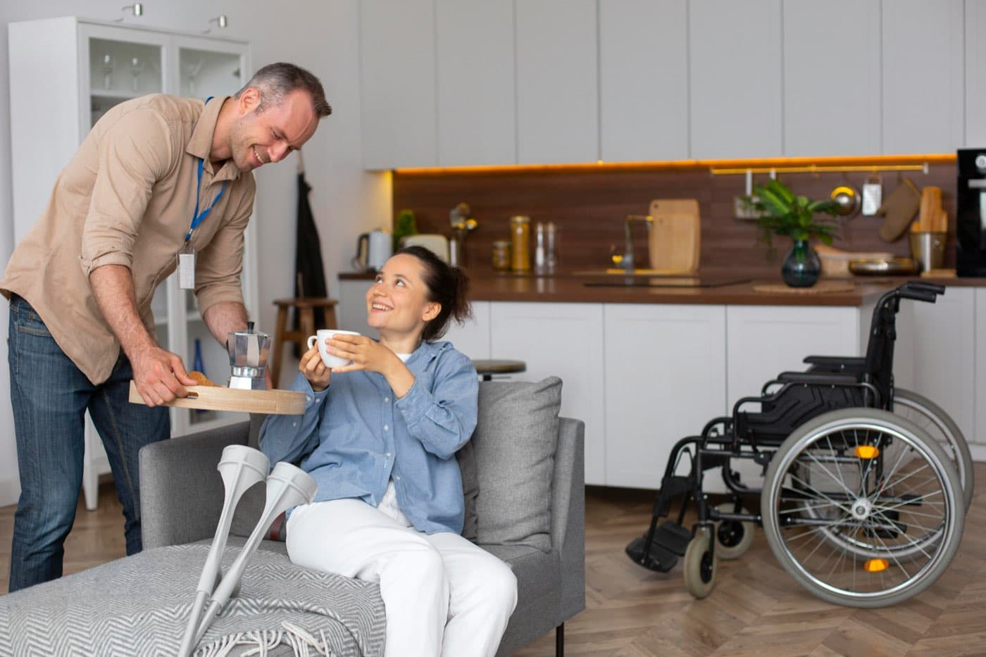 A smiling caregiver bringing a tray of coffee to a woman seated on a sofa in a bright kitchen, with crutches and a wheelchair nearby